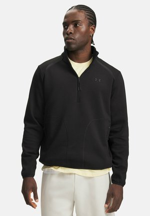 Man met gevlochten haar, die een zwarte sweater met een kwart-ritssluiting draagt over een lichtgeel overhemd en witte broek, kijkt naar de zijkant.