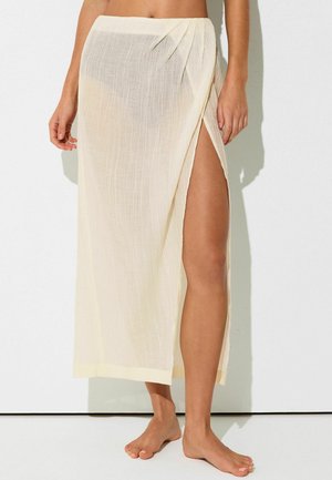 Világos bézs áttetsző anyagú, magas oldalsliccsel rendelkező összehajtott szoknya, mezítláb viseli egy fehér háttér előtt álló modell.