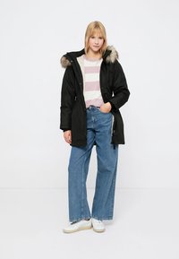 Parka neagră cu glugă din blană sintetică, închidere cu fermoar și buzunare laterale. Purtată peste un pulover alb cu dungi roz deschis și asortată cu blugi largi de culoare albastră.