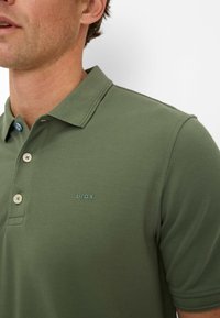 Olive green polo shirt with a textured fabric, two buttons, and a blue accent button. Small "Brax" logo in matching color on the chest.