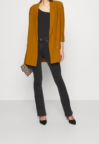 Blazer de doble botonadura mostaza sobre una camiseta sin mangas negra, combinado con jeans acampanados negros y tacones negros de punta. Lleva un bolso de mano impreso.