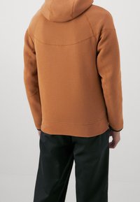 Sweatshirt à capuche marron avec une finition texturée, présentant un détail de couture le long des épaules, associé à un pantalon noir.