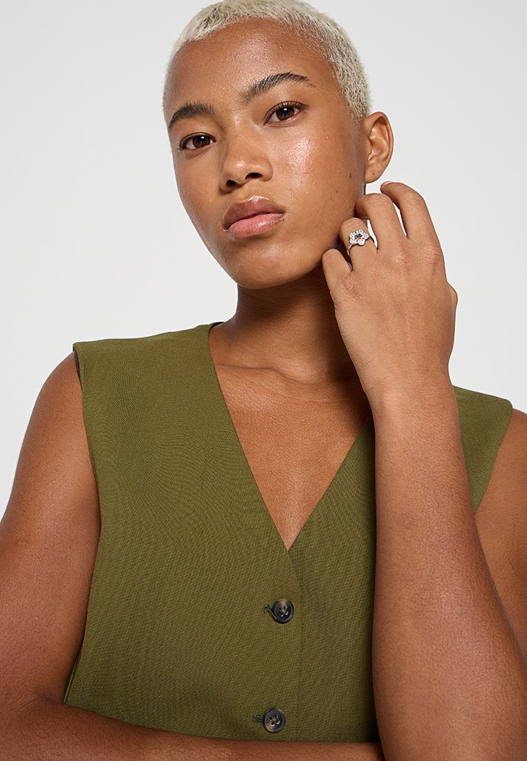 Persona con cabello rubio corto que lleva una blusa sin mangas de color verde oliva, mostrando un gran anillo decorativo en la mano izquierda cerca del rostro.