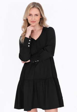 Lyshåret kvinde med bølget hår iført en sort kjole med lange ærmer og hvide knapper på manchetterne, poserer med hånden hvilende på hagen mod en hvid baggrund.