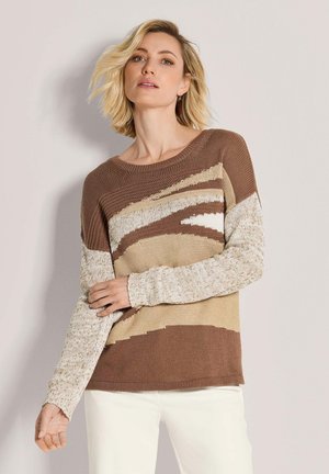 Blondine die een bruin, beige en crème gebreide trui met patroon draagt en een witte broek, staand tegen een effen lichte muur.