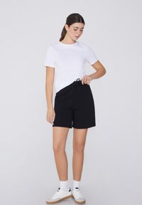Zwarte shorts met een trekkoord in de taille, gecombineerd met een eenvoudige witte T-shirt met korte mouwen. Het model draagt witte sneakers en enkelsokken.