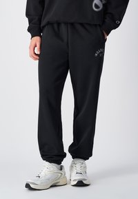 Zwarte joggingbroek met een elastische tailleband en boorden, met een logo-opdruk op de bovenbeen. Het materiaal lijkt zacht en gestructureerd.