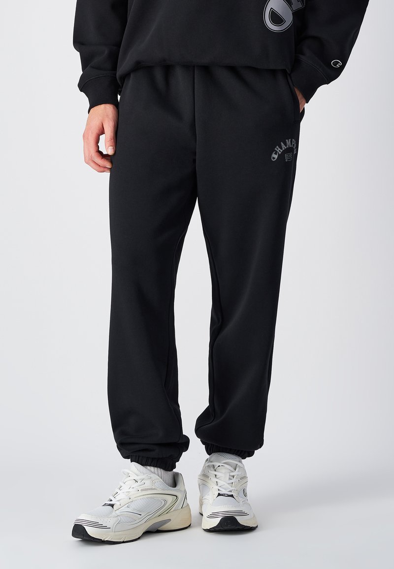 Zwarte joggingbroek met een elastische tailleband en boorden, met een logo-opdruk op de bovenbeen. Het materiaal lijkt zacht en gestructureerd.