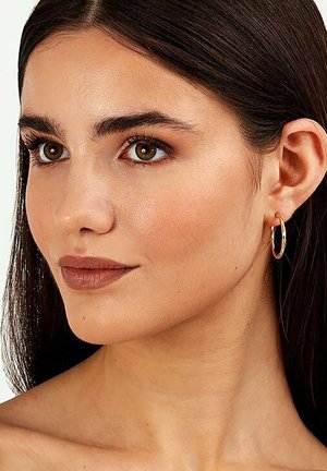 Runder goldener Creolenohrring, der am Ohr getragen wird, mit einer glatten metallischen Oberfläche und einem sicheren Verschluss, kombiniert mit natürlichem Make-up und dunklem Haar.