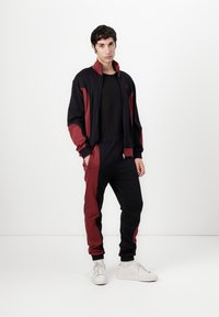 Zwart en bordeaux rood trainingspak van gladde stof, met een ritsjack met zijzakken, elastische manchetten en bijpassende broek.