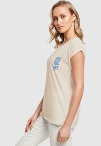 Beige Baumwollt-Shirt mit kurz gerollten Ärmeln und blauem Text "GOOD VIBES ONLY", kombiniert mit engen weißen Hosen.