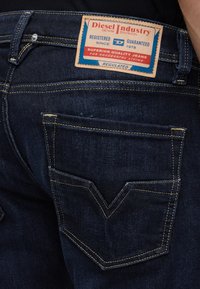 Πίσω τσέπη σκούρου μπλε τζιν με κίτρινα ράμματα και ορθογώνια ετικέτα Diesel Industry πάνω από την τσέπη στη μέση.
