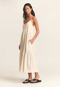 Maxi dress in light beige fabric, featuring a V-neckline, thin straps, side pockets, and a tiered hemline. Worn with black sandals.