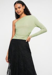 Top verde-claro de manga longa com um ombro exposto, combinado com uma saia preta rendada com um design fluido e tecido texturizado.
