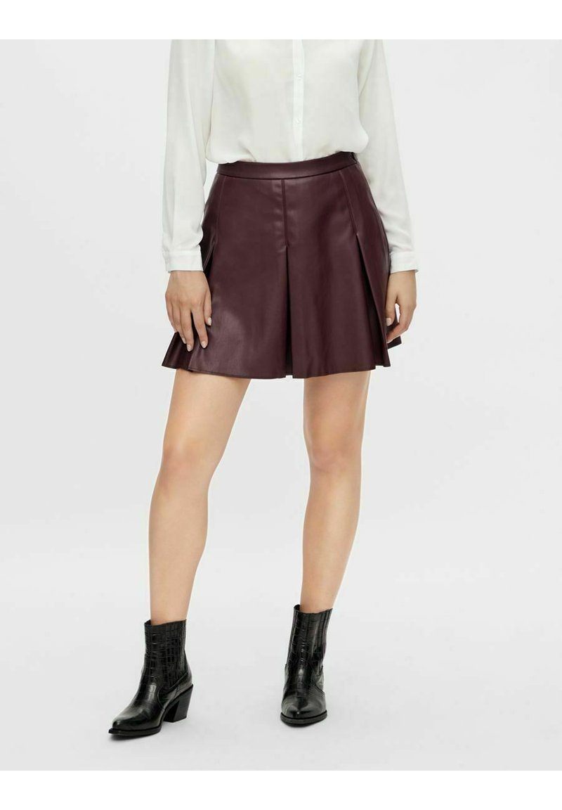 Jupe en cuir bordeaux avec des plis et une taille cintrée, associée à des bottines en croco noires et à une chemise blanche à boutons.