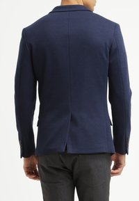 Marinblå blazer i textil med struktur, med spetsiga slag, en enda bakventil och knapplister på ärmarna. Skräddarsydd passform.