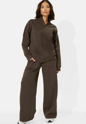 Donna in piedi con una mano in tasca, indossa una felpa marrone con zip e pantaloni a gamba larga con sneakers bianche su sfondo bianco.