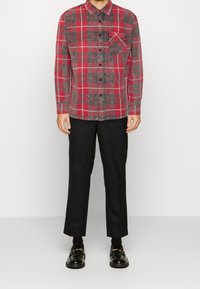 Camicia di flanella a quadri rossi e grigi con maniche lunghe, colletto con bottoni e taschino sul petto; abbinata a pantaloni neri e scarpe.