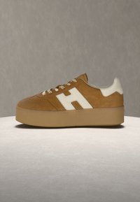 Zapatilla de ante marrón con suela gruesa de goma, detalle blanco en el talón, cordones blancos y un gran logo "H" blanco en el lateral, sobre un fondo neutro.