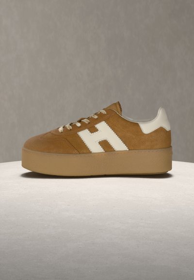 Brązowy zamszowy trampek z grubą gumową podeszwą, białym akcentem na pięcie, białymi sznurowadłami i dużym białym logo "H" po boku, na neutralnym tle.