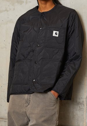 Person iført en sort quiltet jakke med trykknapper og tre forlommer, hvoraf den ene har et Carhartt-logo patch.