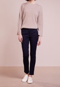 Pull en maille beige avec des manches larges, associé à un pantalon slim-fit navy et des baskets blanches, sur un fond rose doux.