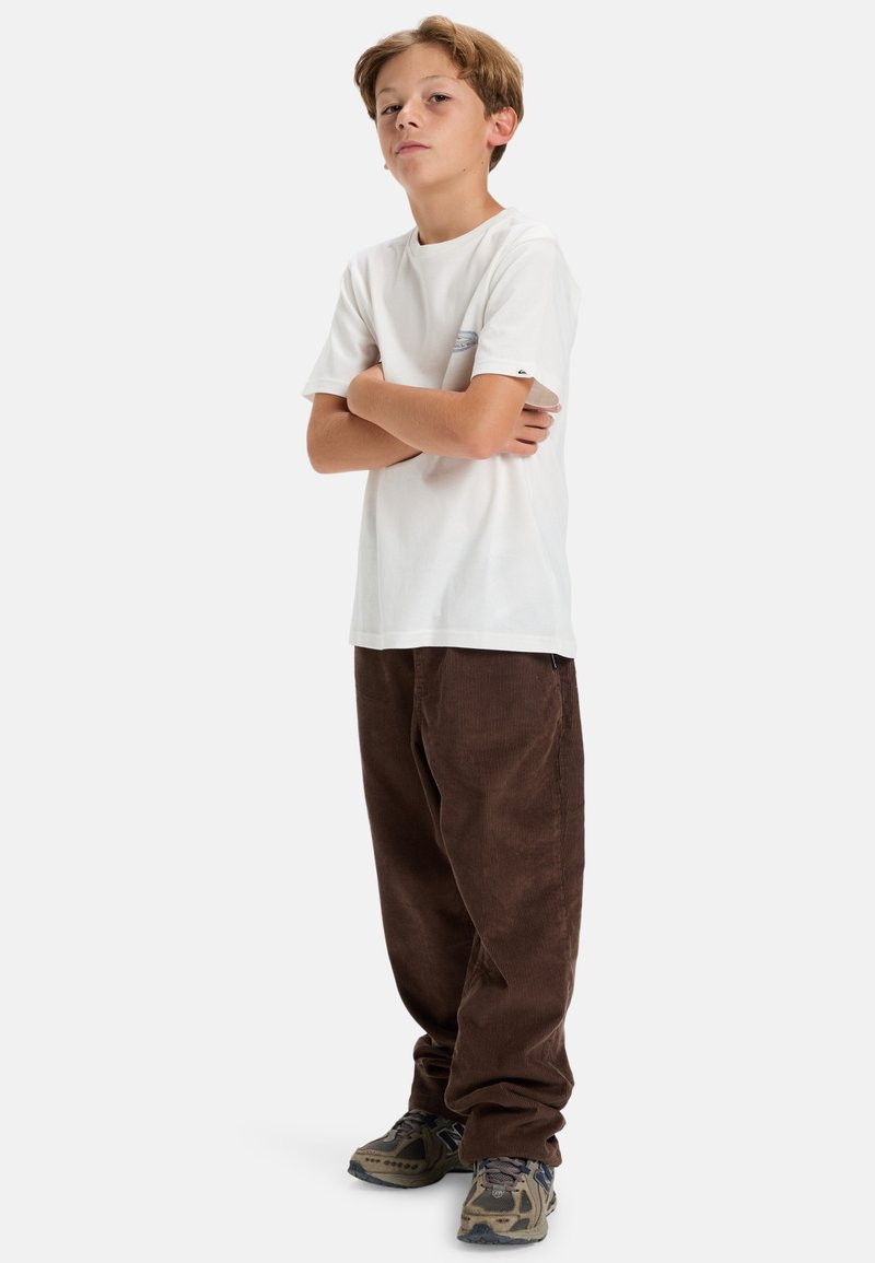 Hvit T-skjorte med korte ermer og blå logo, kombinert med brune kordfløyelsbukser. Modellen har på seg grå og svarte mønstrede joggesko.