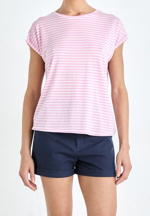 Persoon draagt een roze-wit gestreept shirt met korte mouwen en marineblauwe shorts met opgerolde zoom, staand tegen een witte achtergrond.