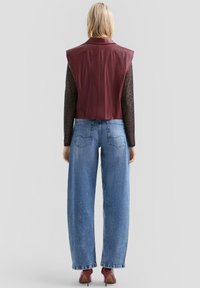 Frau steht mit dem Rücken zur Kamera und trägt eine kastanienbraune Leder-Sattelweste ohne Ärmel, ein durchscheinendes schwarzes Tiermuster-Oberteil, lockere blaue Jeans und kastanienbraune High Heels.
