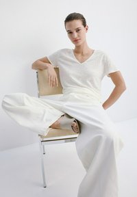 Jeune personne assise sur une chaise, une jambe croisée, portant un t-shirt blanc à col en V, un pantalon blanc à jambes larges et des sandales beiges sur un fond blanc.