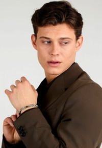 Blazer marrone indossato sopra una camicia scura, con un bracciale intrecciato argento e nero al polso. Texture liscia con una leggera lucentezza.