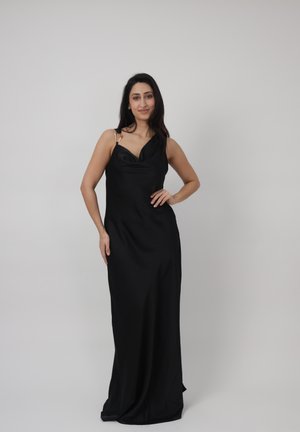 Frau mit langen dunklen Haaren, die ein schwarzes, ärmelloses Abendkleid trägt und mit einer Hand in der Hüfte vor einem schlichten hellen Hintergrund steht.