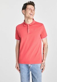 Korallenowa koszulka polo z kołnierzykiem, trzema guzikami i krótkimi rękawami, zdobiona jasnoniebieskimi akcentami. Noszona z jasnymi dżinsami.