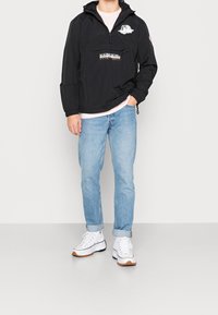 Zwarte pulloverjack met een voorzak, capuchon en logo's; gecombineerd met lichtblauwe spijkerbroeken en witte sneakers met tan accenten.