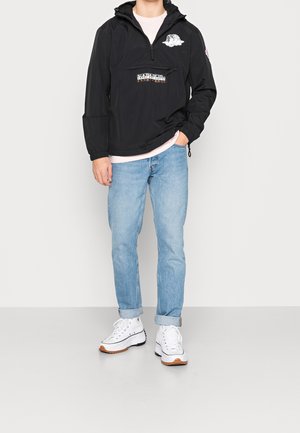 Schwarze Pulloverjacke mit einer Vorderseite-Tasche, Kapuze und Logos; kombiniert mit hellblauen Jeans und weißen Sneakers mit braunen Akzenten.