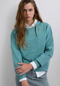 Young woman with long hair wears teal hoodie embossed with skull and crossbones logo, white shirt underneath, and zebra print pants.