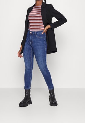 Mujer con un top a rayas rosa y marrón, blazer negro, vaqueros ajustados azules y botas negras gruesas, de pie con una mano en la cadera.