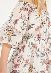 Vestido blanco sin hombros con patrones florales en rosa, amarillo y azul, y mangas con volantes en una persona con cabello largo y rubio.