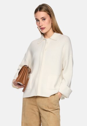 Frau trägt einen weißen Reißverschluss-Pullover und beige Hose, hält eine braune Leder-Clutch vor einem einfarbigen hellen Hintergrund.