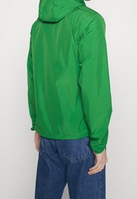 Giacca a capuccio verde con finitura lucida, caratterizzata da una vestibilità comoda e polsini regolabili, abbinata a jeans in denim blu.