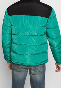 Tealfärgad puffjacka med en svart övre del, med en kviltad textur och elastiska ärmslut. Bärs över blå denimjeans.