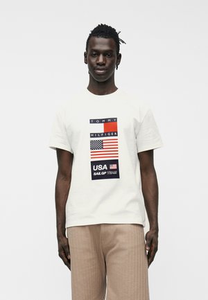 Fiatal férfi sötét bőrrel és rövid dreadlockkal, fehér Tommy Hilfiger pólót visel az USA zászlójával és bézs nadrágot, egy egyszínű háttér előtt áll.