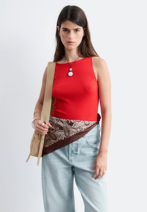 Femme portant un top rouge sans manches, un jean bleu clair, une ceinture écharpe paisley marron, un sac à main beige et un collier pendentif sur un fond uni.