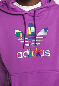 Lila hoodie med en stor, flerfärgad Adidas trefoil-logotyp och varumärkesnamnet "adidas" i fetstil, färgglada bokstäver.