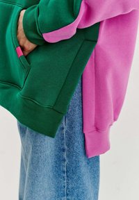 Mikina má zelené telo a ružové rukávy, vyrobená z mäkkého materiálu. Obsahuje prednú kapsu a rebrované manžety. Nosená s svetlomodrými džínami.