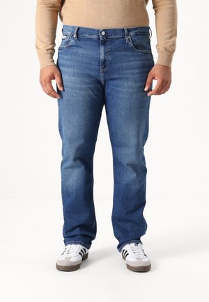 Personne portant un jean droit bleu, un haut beige à manches longues et des baskets blanches avec des bandes noires, debout devant un fond blanc.