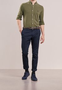 Camicia verde a maniche lunghe con vestibilità slim, pantaloni blu scuro, cintura marrone intrecciata e scarpe da ginnastica scure. Tessuto morbido, linee pulite e maniche arrotolate.