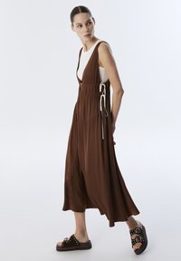 Bruine mouwloze jumpsuit met gathers bij de taille en zijstrikjes, gecombineerd met zwarte plateausandalen met gestikte accenten.