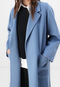 Jasnoniebieski wełniany płaszcz o luźnym kroju, z klapami z nacięciem i dwoma bocznymi kieszeniami, założony na czarny sweter i białą koszulę.