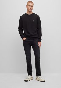 Giovane uomo in piedi con una mano in tasca, indossa un felpa nera, jeans neri e sneakers bianche contro uno sfondo grigio uniforme.